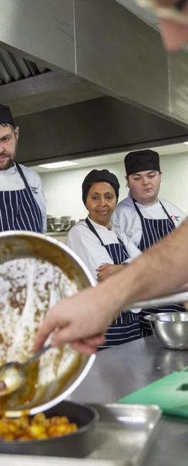A group of culinary students in a professional kitchen observe closely as their instructor pours a sauce from a pan into a small dish. A group of culinary students in a professional kitchen observe closely as their instructor pours a sauce from a pan into a small dish.